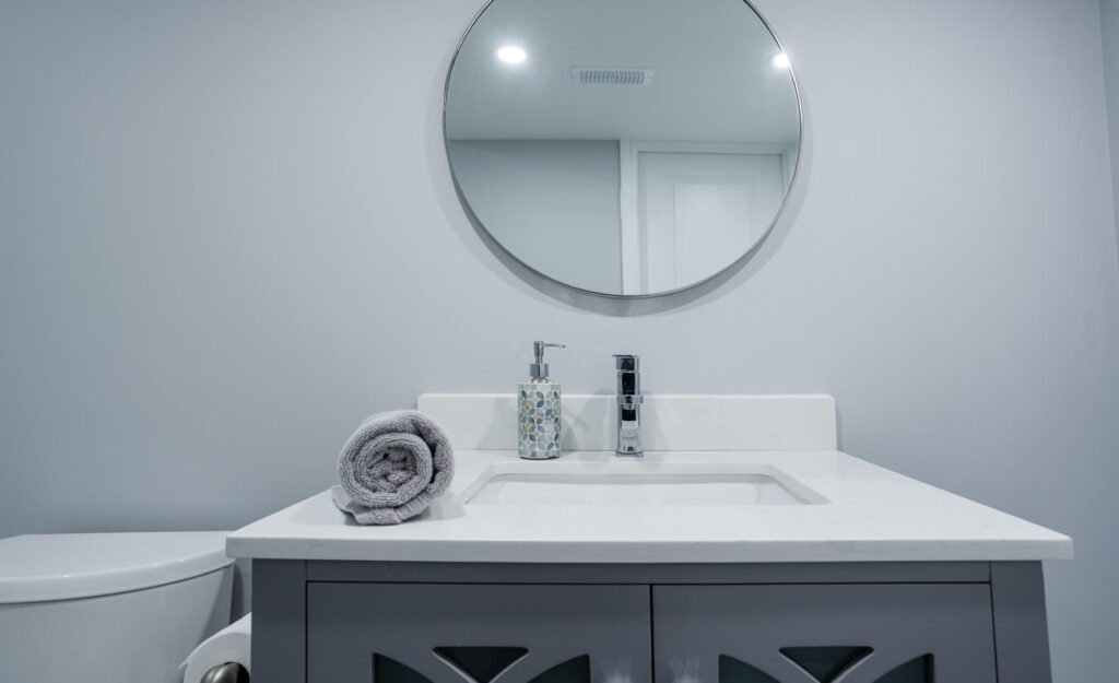 contemporary basement bathroom with round mirror and white ceramic countertop with vanity